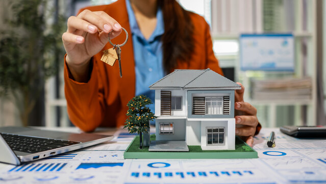 Real estate agent holding house keys beside model home on desk with financial charts and laptop, property purchase, mortgage and investment concept, close-up, shallow depth of field