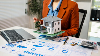 Businesswoman holding model house above desk with laptop, charts, calculator, keys in modern office, real estate finance and mortgage planning concept © Phushutter