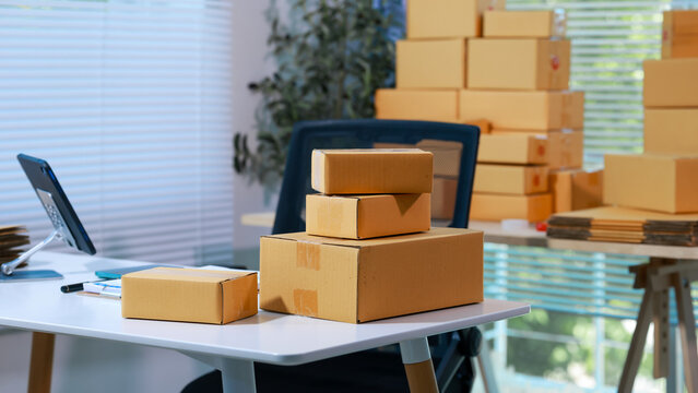 Office workspace with stacks of cardboard shipping boxes on desk and in background, laptop and supplies, daylight through blinds; ecommerce fulfillment, moving or storage concept