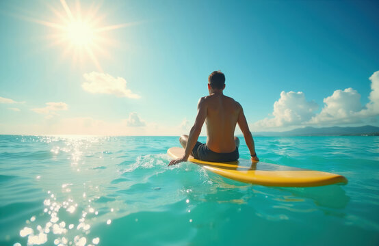 Man sits on yellow surfboard in blue ocean water. Bright sun shines on calm sea surface. Peaceful summer day at seaside. He waits for waves.