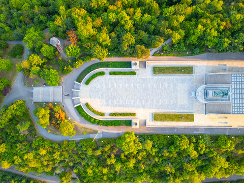 The National Monument at Vitkov is seen at sunrise. The area is surrounded by trees, and the Tomb of the Unknown Soldier is visible. People can be seen walking and enjoying the morning.