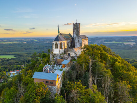 Bezdez Castle stands proudly atop a hill as the sun sets in the background. The medieval ruins are surrounded by lush greenery, offering a stunning view during the summer evening.