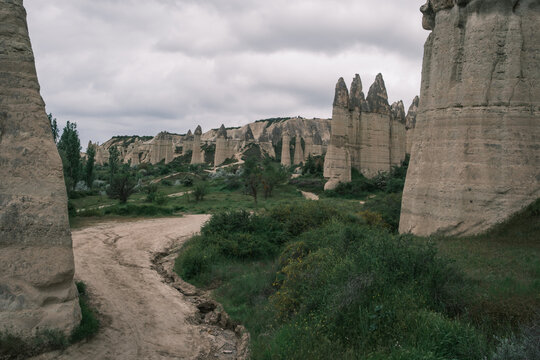 Dirt trail winding through the iconic rock pillars of Love Valley in Cappadocia, Turkey. Unique volcanic tuff formations surrounded by green vegetation under an overcast sky.