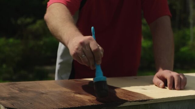 Handmade furniture creation, repair, restoration. Man staining wooden board with brown paint outdoors