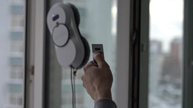 Robotic window cleaner with rotating microfiber pads washing glass surface. Closeup person hand holding remote control, guiding smart device for convenient household chore automation, slow motion.