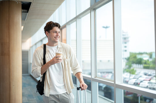 Happy white caucasian man tourist holding coffee cup and luggage while standing or waiting in airport
