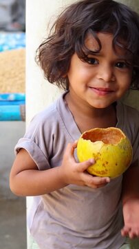 Cute Indian toddler girl drinking fresh wood apple juice directly from its natural shell, happy village child enjoying healthy traditional bel sharbat, authentic rural lifestyle
