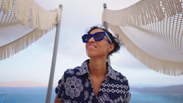 Stylish Indian woman in blue sunglasses and patterned shirt smiles at luxury resort terrace in Santorini Greece with ocean views and decorative hammock canopies swaying in the breeze at dusk.