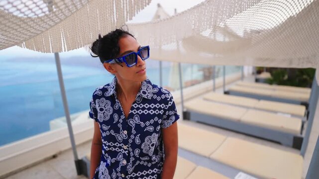 Stylish Indian woman wearing blue sunglasses and patterned shirt stands at luxury resort terrace in Santorini Greece. Sun loungers and white umbrellas frame the stunning sea view backdrop.