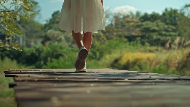 Low angle rear view of woman in white dress and sneakers walking on wooden path at park during sunny day carefree female traveler enjoying nature journey slow living and mindfulness concept