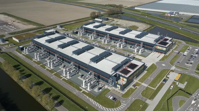 Large scale drone footage of a modern cloud computing facility surrounded by canals and green fields in the Netherlands.