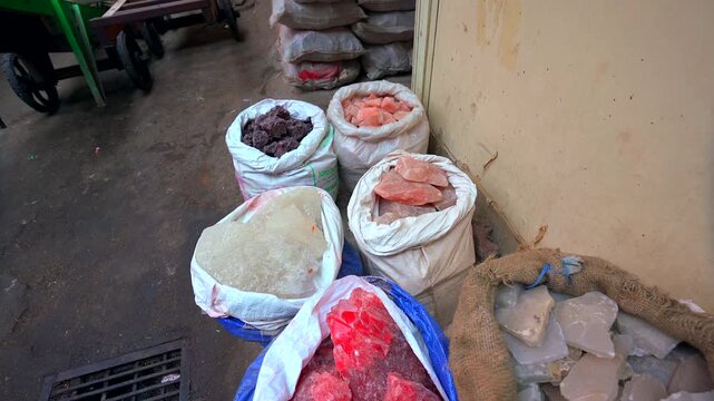 A high-angle static shot of traditional woven sacks filled with large raw blocks of pink Himalayan salt, white alum, and natural mineral crystals at a market stall in India