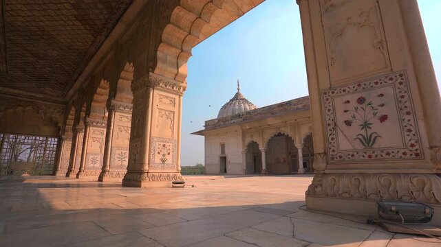 A low-angle static shot from inside an intricately carved pavilion, framing a beautiful white marble dome structure under a clear sky at the historic Red Fort in Delhi, India.