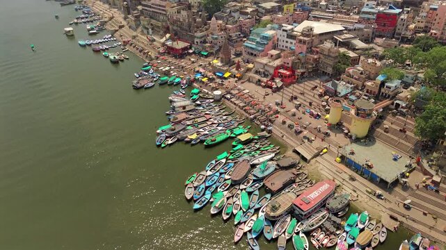 An aerial wide static shot showcasing the historic Dashashwamedh Ghat with rows of colorful tourist boats and an ancient stone Hindu temple on the Ganges River in Varanasi, India