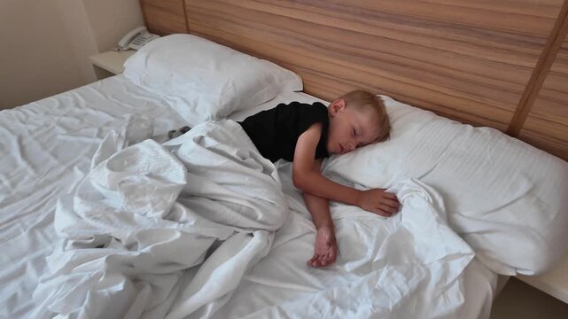 High angle view of a little boy sleeping alone in the middle of a large double bed with white crumpled sheets in a hotel room.