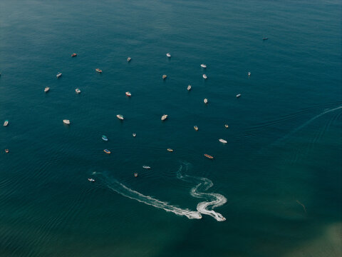 Aerial view of Baga Beach with numerous small boats anchored in the turquoise sea and jet skis leaving white wake trails on the water surface in Baga, Goa, India.