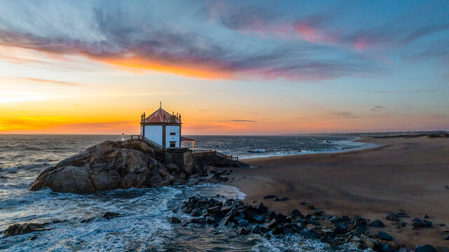 Aerial view of the Capela do Senhor da Pedra chapel built on a rocky outcrop at sunset with waves crashing on the beach in Miramar, Porto, Portugal.