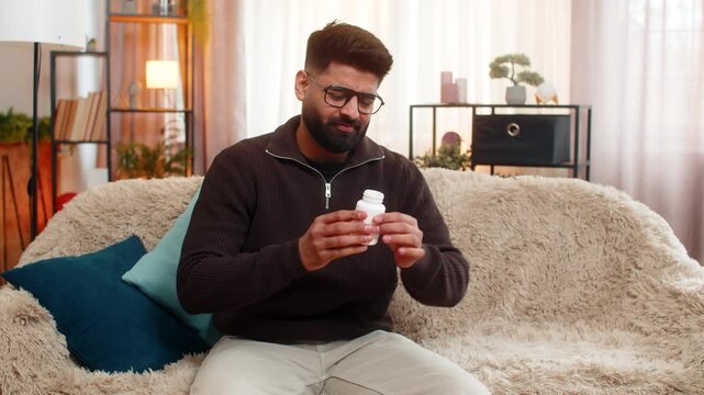 Indian man at home holds stomach with painful expression, sitting on sofa while feeling nausea. Arabian guy struggles with food poisoning or stomach trouble, showing clear signs of digestive pain