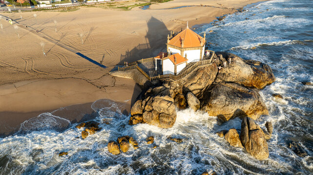 Aerial view of Capela do Senhor da Pedra on a rocky outcrop with waves crashing and long shadows on the sandy beach in Miramar, Porto, Portugal.