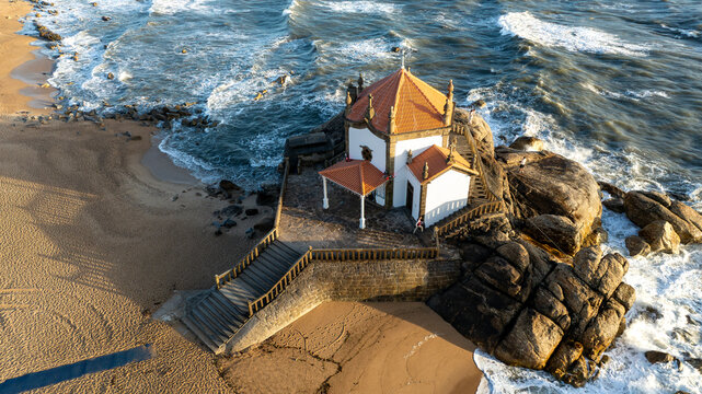 Aerial view of Capela do Senhor da Pedra built on a rocky outcrop with people on the stairs surrounded by crashing waves and a sandy beach in Miramar, Porto, Portugal.