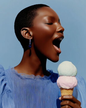 Black woman with shaved head, blue drop earring and open mouth eating a double scoop mint and strawberry pink ice cream waffle cone wearing a blue chiffon top, joyful fashion editorial concept