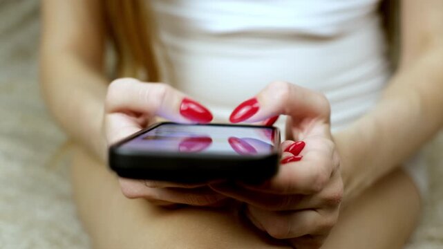 Young woman with red manicure using a smartphone, texting messages and scrolling on a social network feed, close up shot