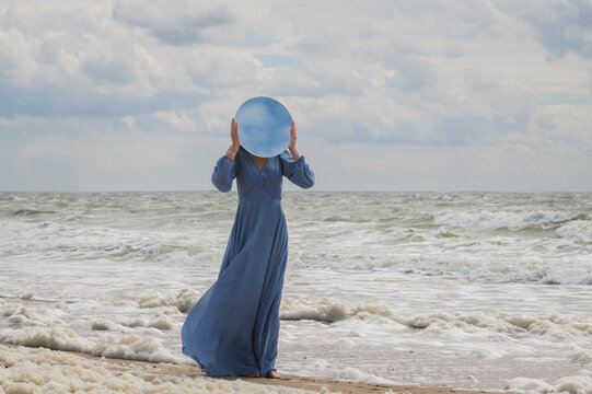 art portrait of woman in blue dress hiding face behind round mirror on beach near ocean