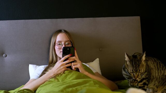 Cheerful young woman texting on her smartphone in bed, while her adorable tabby cat walks on the blanket seeking attention and cuddles