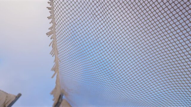 Close-up view of woven mesh net canopy with decorative fringe edges stretching across frame against soft blue sky, showing intricate diamond pattern texture and gentle movement in Mediterranean.