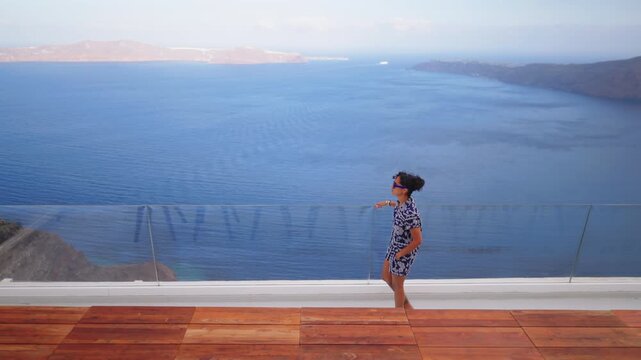 Stylish Indian woman in blue floral dress walks barefoot across luxury wooden deck terrace with modern glass railing overlooking stunning Santorini caldera.