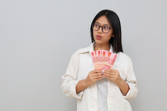 Photo of a young Asian woman wearing casual clothes holding Indonesian rupiah banknotes, looking to the side while contemplating something, standing isolated on a white background.