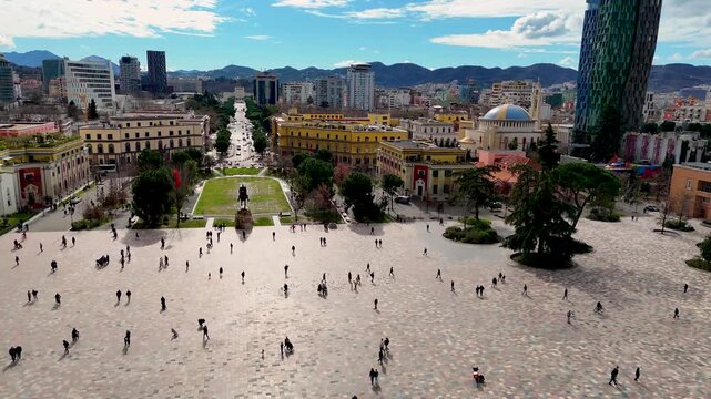 Aerial perspective rising above Skanderbeg Square in Tirana, Albania, revealing pedestrian crowds, the Skanderbeg Monument, historic architecture, and distant mountain peaks