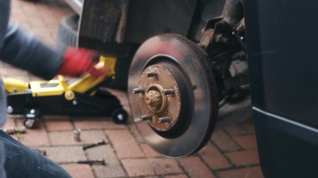 Worker hammering wooden block against brake disc for removal. Automotive repair loosening seized brake rotor from wheel hub during car maintenance. Closeup manual disassembly with tools during vehicle