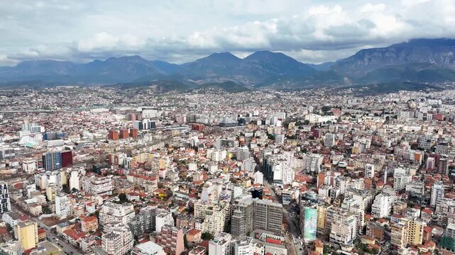 High-angle aerial perspective pulling away and flying backward over Tirana, Albania, revealing an expansive urban landscape, residential blocks, and a majestic mountain range backdrop