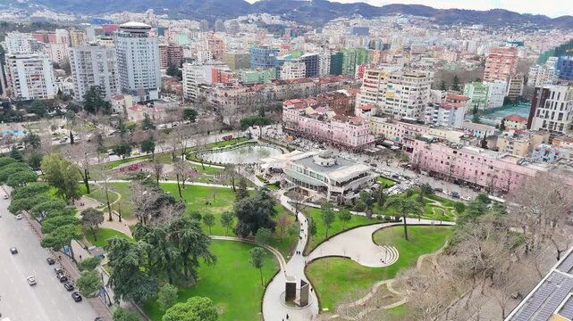 Aerial perspective flying forward over Rinia Park in central Tirana, Albania, panning down on the green lawn pathways, trees, city streets, and the central park water fountain