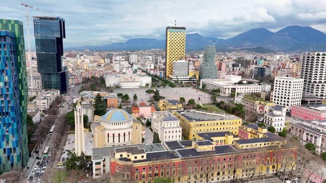 Aerial view flying forward and moving in toward Skanderbeg Square in Tirana, Albania, showcasing the Resurrection Cathedral, modern skyscrapers, and a mountain horizon