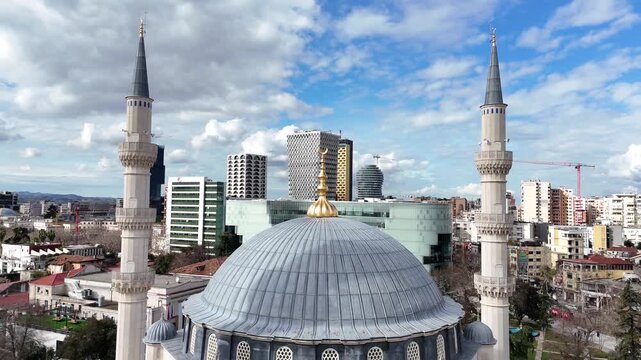 Aerial camera descends vertically behind the central blue-grey dome, framing two flanking minarets and revealing the modern downtown skyline of Tirana, Albania