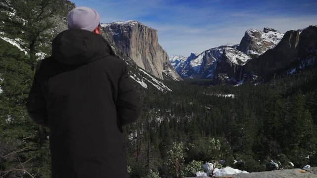 Solo hiker overlooking yosemite valley view in winter, backfacing traveler in black parka and beanie gazing toward El Capitan, snow patches on pine slopes, rocky foreground and reflective