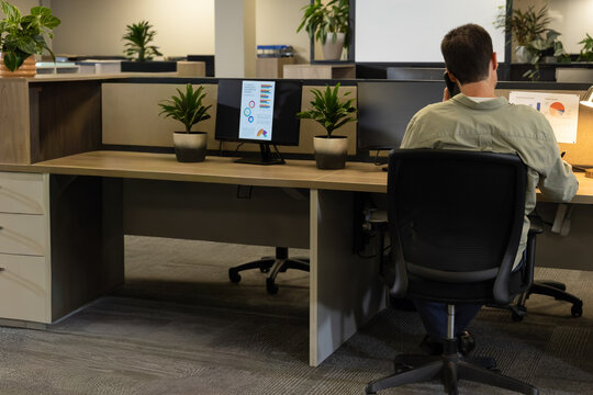 Mature man sitting in ergonomic chair at open office holding phone two monitors chart, copy space