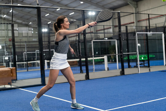 Mature female playing padel, reaching with padel racket for ball on blue court inside hall