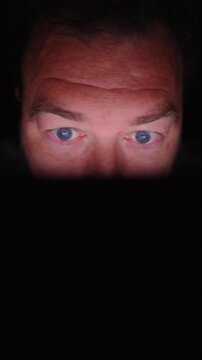 Vertical Screen: Close-up of middle-aged man with piercing blue eyes illuminated by soft screen glow in complete darkness, face partially visible with intense focused gaze and furrowed brow