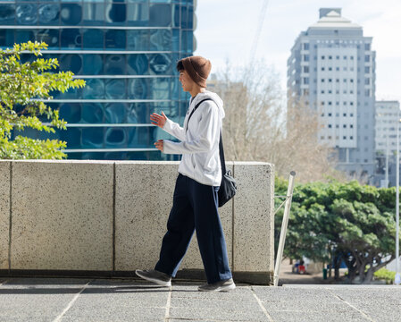 Asian adult man walking along tiled plaza behind low parapet wearing beanie carrying black bag