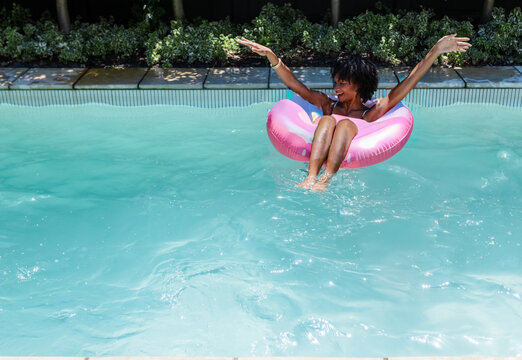 African American woman floating on pink donut float in blue pool wearing bikini, copy space