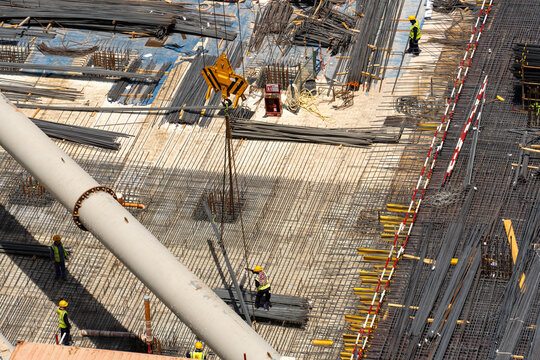 Aerial view of a busy construction site with workers in safety gear laying steel rebar near a large crane hook and industrial piping in Dubai, Dubai, United Arab Emirates.