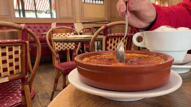 Woman's hand breaking the caramelized sugar crust of a traditional French cr&egrave;me br&ucirc;l&eacute;e with a spoon in a classic bistro in Paris, enjoying a sweet dessert with a hot cappuccino