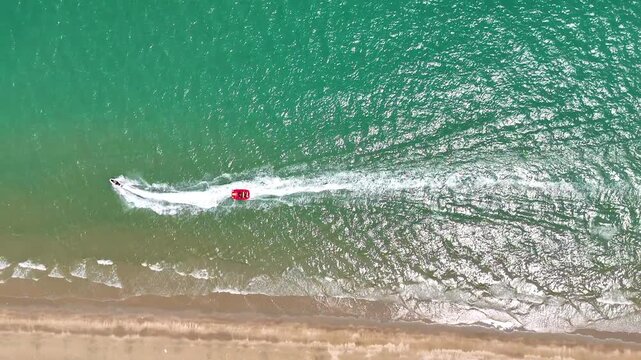 Drone footage aerial view of a fast jet ski pulling a red inflatable banana boat with tourists on turquoise sea water near the coast