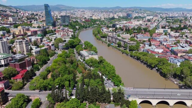 Wide aerial panorama of the Kura River (Mtkvari) flowing through central Tbilisi, Georgia, featuring the Galaktion Tabidze Bridge, lush riverbank parks, and the modern Biltmore Hotel skyscraper
