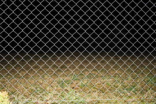 ​A metal chain-link fence is installed against the background of illuminated dry grass and deep night darkness. The clear geometric structure of the fence creates a distinct diamond pattern in the fla