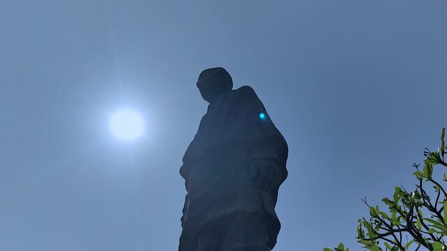 Low-angle silhouette of Statue of Unity with blazing sun flare and tropical tree branches framing deep blue sky. Ideal for cinematic openers, India heritage, and patriotic content.
