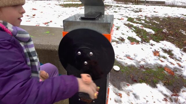 Side view tracking shot of a young child turning a kinetic hand crank wheel at an interactive park science game display. Snow covers the playground lawn at Varosliget city park in Budapest Hungary.